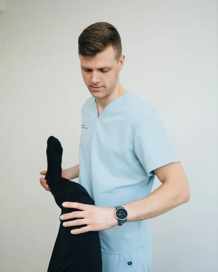 Owen Castellano in clinic scrubs, guiding a patient’s leg through a stretch.