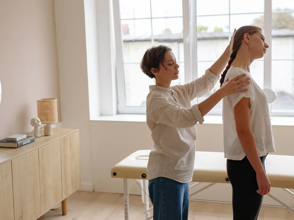A therapist adjusts a standing patient’s head position in a bright room.