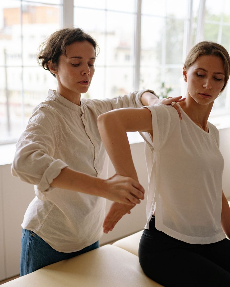 A therapist guides a patient through an arm stretch on a treatment table beside a bright window.